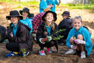 Picture of three crouched girls holding tree seedlings and smiling, while other children are planting trees in the background