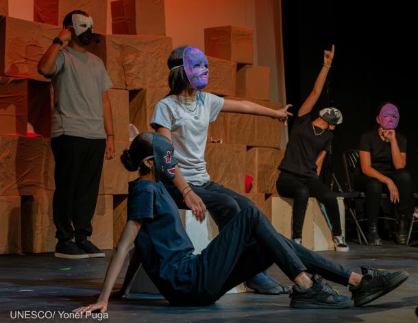 Personas jóvenes usando máscaras apuntan a diferentes lugares. Se trata de una obra de teatro. 