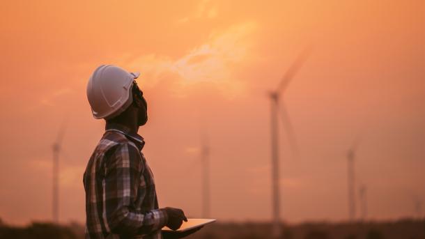 African engineer man windmills wearing white hard hat and using tablet with wind turbine on silhouette sunset