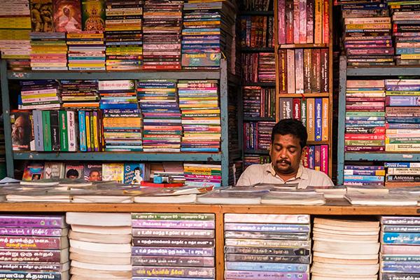 A book seller sitting in a shop with stacks of academic books on the shelves. Tamil Nadu, India